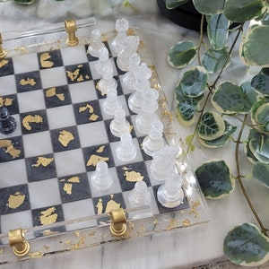 May include: A clear acrylic chessboard with gold leaf accents. The board has black and white squares and comes with black and white chess pieces.
