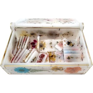 Puede incluir: Una caja blanca con un borde dorado y un dise&ntilde;o floral en la parte inferior. La caja est&aacute; abierta y llena de fichas de domin&oacute; blancas con flores prensadas y puntos dorados.