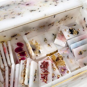 Puede incluir: Una caja blanca con ribete dorado, llena de fichas de domin&oacute; hechas a mano. Las fichas de domin&oacute; est&aacute;n hechas de resina transparente con flores prensadas y detalles dorados.