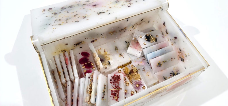 Puede incluir: Una caja blanca con ribete dorado, llena de fichas de domin&oacute; hechas a mano. Las fichas de domin&oacute; est&aacute;n hechas de resina transparente con flores prensadas y detalles dorados.