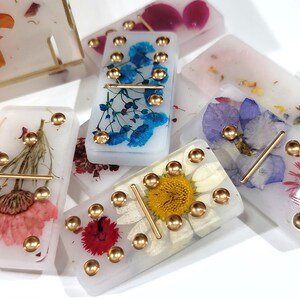 Puede incluir: Un juego de domin&oacute; blanco con puntos dorados y flores prensadas. Cada domino tiene un dise&ntilde;o floral diferente, incluyendo flores azules, rosas y amarillas.