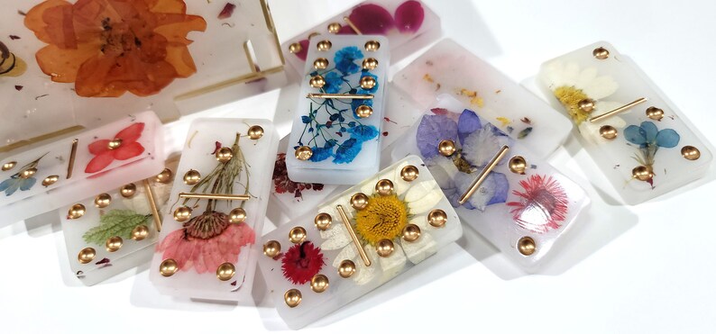 Puede incluir: Un juego de domin&oacute; blanco con puntos dorados y flores prensadas. Cada domino tiene un dise&ntilde;o floral diferente, incluyendo flores azules, rosas y amarillas.