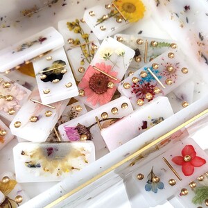 Puede incluir: Un juego de domin&oacute; blanco con flores prensadas y detalles dorados. Los domin&oacute;s est&aacute;n en una caja transparente con un borde dorado.