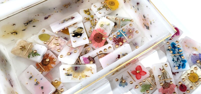 Puede incluir: Un juego de domin&oacute; blanco con flores prensadas y detalles dorados. Los domin&oacute;s est&aacute;n en una caja transparente con un borde dorado.