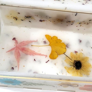 Puede incluir: Una caja blanca con un borde dorado y una tapa transparente. El interior de la caja est&aacute; lleno de flores y hojas prensadas, incluyendo una hoja de arce rosa, una hoja de ginkgo amarilla y una flor amarilla.