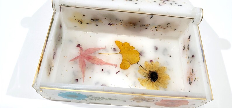 Puede incluir: Una caja blanca con un borde dorado y una tapa transparente. El interior de la caja est&aacute; lleno de flores y hojas prensadas, incluyendo una hoja de arce rosa, una hoja de ginkgo amarilla y una flor amarilla.