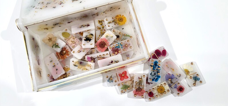 Puede incluir: Un juego de domin&oacute; blanco con flores prensadas y detalles dorados. Los domin&oacute;s est&aacute;n en una caja blanca con un borde dorado.