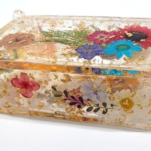 Puede incluir: Una caja rectangular transparente con tapa, llena de flores prensadas y pan de oro. Las flores son en tonos de rosa, morado, azul y amarillo.