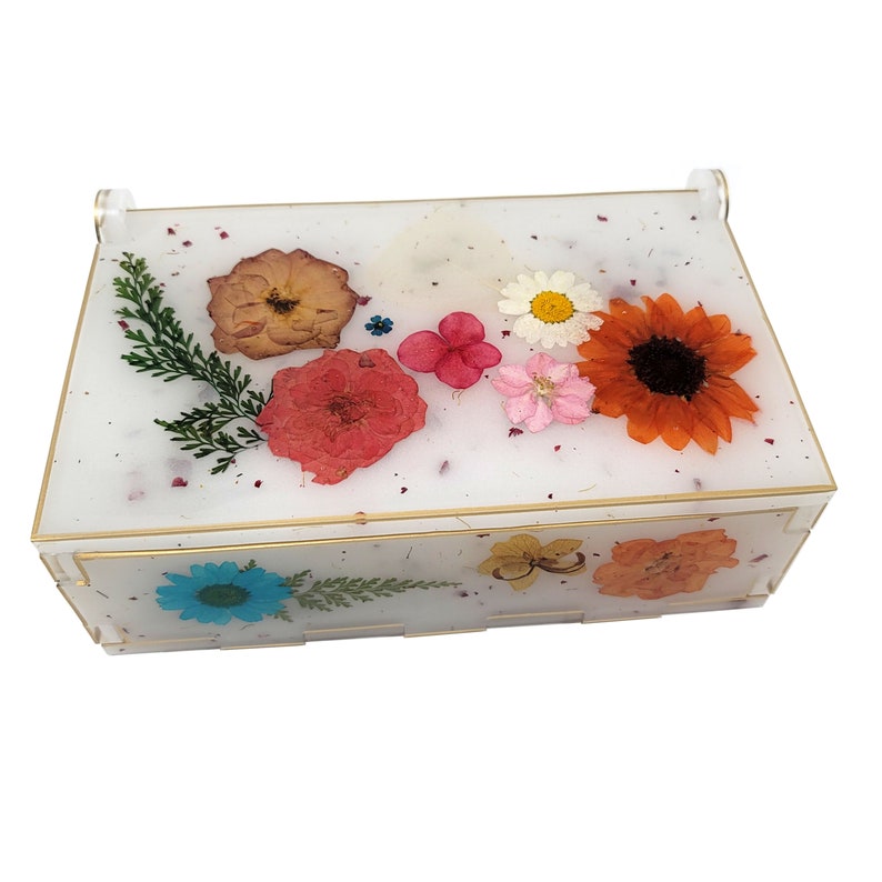 Puede incluir: Una caja rectangular blanca con un borde dorado, llena de flores y hojas prensadas. Las flores son en tonos de rosa, rojo, naranja, amarillo y azul.