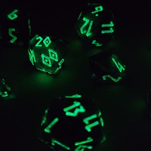 Puede incluir: Un conjunto de ocho dados que brillan en la oscuridad con números verdes. Los dados están hechos de un material translúcido y se iluminan con una luz negra.