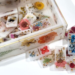 Puede incluir: Un juego de domin&oacute; blanco con detalles dorados y flores prensadas. Los domin&oacute;s est&aacute;n en una caja blanca con un borde dorado y flores prensadas.