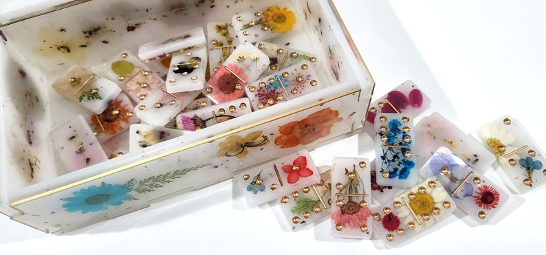 Puede incluir: Un juego de domin&oacute; blanco con detalles dorados y flores prensadas. Los domin&oacute;s est&aacute;n en una caja blanca con un borde dorado y flores prensadas.