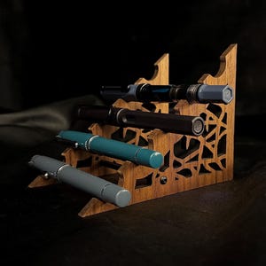May include: A wooden pen display stand with four pens. The stand is made of light brown wood with decorative cutouts. The pens are various colors, including gray, teal, and black, with silver accents. The background is dark.