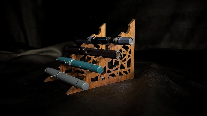 May include: A wooden pen display stand with four pens. The stand is made of light brown wood with decorative cutouts. The pens are various colors, including gray, teal, and black, with silver accents. The background is dark.