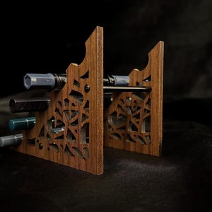May include: A wooden pen holder with a decorative, geometric cut-out design. The holder is made of a warm brown wood and has slots to hold several pens. The pens are various colors, including gray, black, and teal.