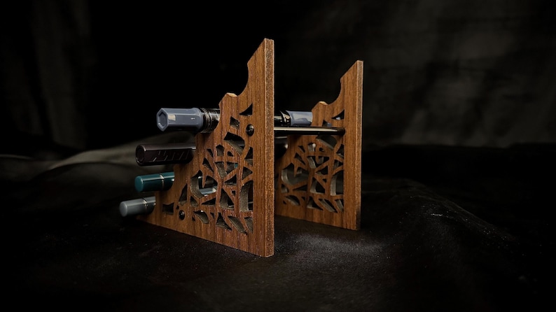 May include: A wooden pen holder with a decorative, geometric cut-out design. The holder is made of a warm brown wood and has slots to hold several pens. The pens are various colors, including gray, black, and teal.