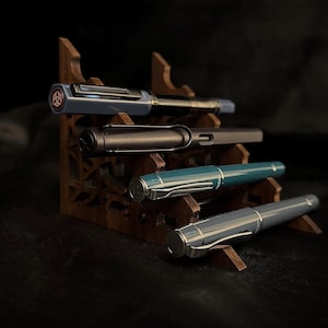 May include: Four fountain pens in various colors, including blue, black, teal, and gray, are displayed on a wooden pen stand. The pens have silver accents and are arranged on different levels of the stand, showcasing their design.