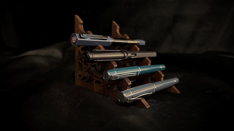 May include: Four fountain pens in various colors, including blue, black, teal, and gray, are displayed on a wooden pen stand. The pens have silver accents and are arranged on different levels of the stand, showcasing their design.