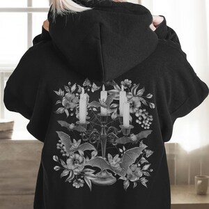 May include: Black hoodie featuring a graphic print of a candelabra with three candles, bats, and flowers in shades of grey.