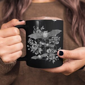 May include: Black ceramic mug with a white design of bats and flowers. The bats are flying in a circle around a spiderweb.