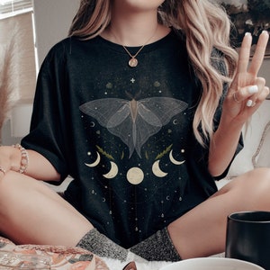 以下が含まれることがあります： 白い蛾と三日月のデザインが施された黒いTシャツ。ゆったりとしたフィット感とクルーネックです。