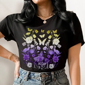 Op de afbeelding: Zwart T-shirt met een grafische print van paarse en gele bloemen, vlinders en paddenstoelen.