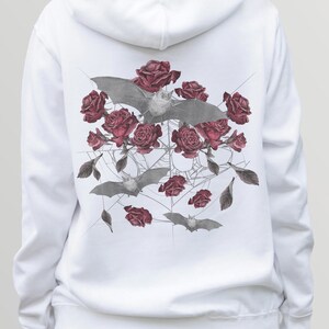 May include: White hoodie featuring a detailed graphic of gray bats, deep red roses, and a spiderweb design. The artwork is centered on the back of the hoodie. The design has a gothic or alternative aesthetic.