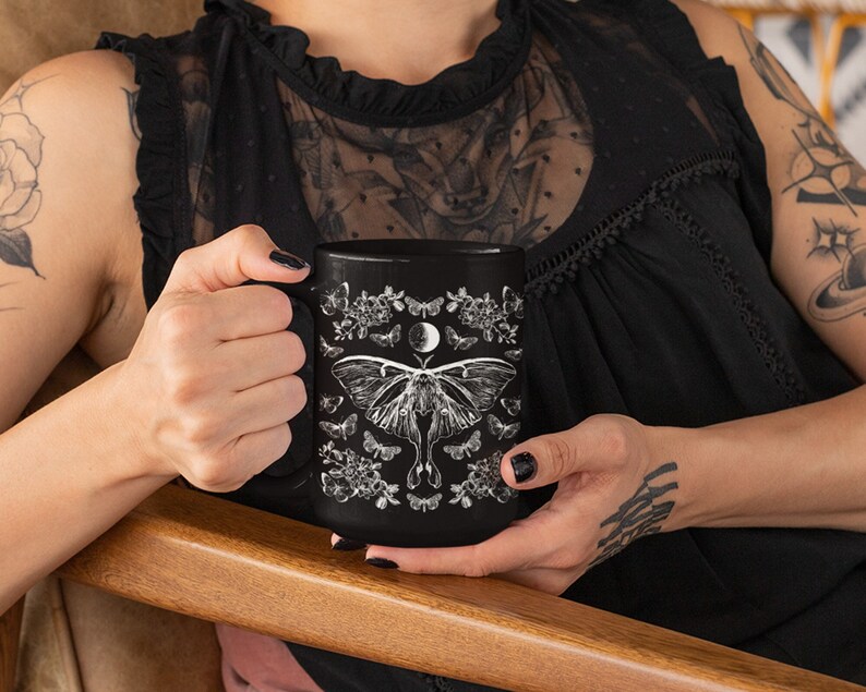 May include: Black ceramic mug featuring a white illustration of a moth, moon, and floral elements. The mug has a large handle and is held by a person. The design is symmetrical and detailed, creating a gothic or celestial theme.