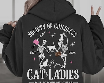 May include: Black hoodie with a white graphic print of two skeletons holding champagne glasses and cats. The text reads "Society of Childless Cat Ladies Est 2024 - R.I.P. to when we gave AF - "