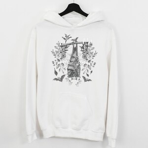 May include: White hoodie featuring a black and white illustration of a bat hanging upside down from a branch with candles and floral accents. The design includes additional bats and botanical elements, creating a gothic aesthetic.