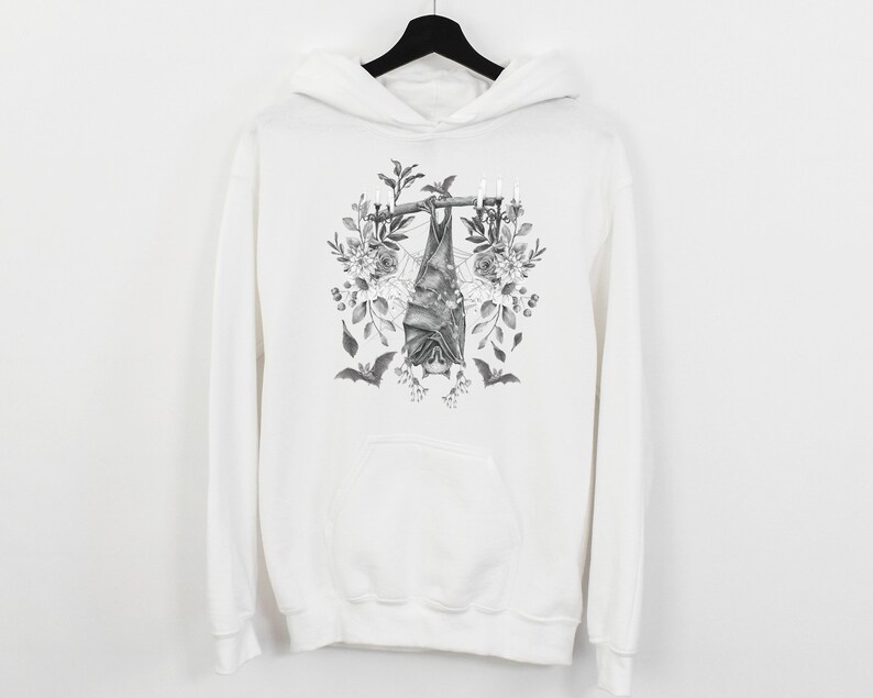 May include: White hoodie featuring a black and white illustration of a bat hanging upside down from a branch with candles and floral accents. The design includes additional bats and botanical elements, creating a gothic aesthetic.