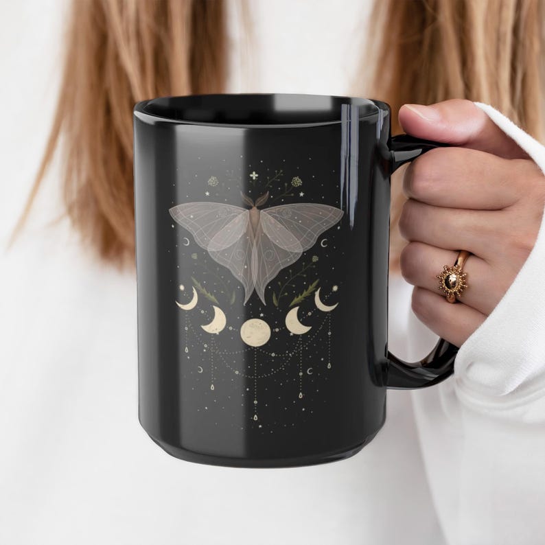 Pode incluir: Caneca de cer&acirc;mica preta com um design de mariposa branca e luas crescentes. A mariposa tem asas transl&uacute;cidas e est&aacute; cercada por pequenas estrelas e elementos florais.