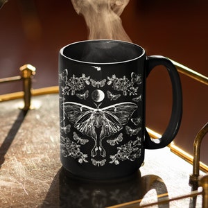 May include: Black ceramic mug featuring a white illustration of a moth, butterflies, flowers, and a crescent moon. The mug has a large handle and is filled with steaming liquid, suggesting a hot beverage.