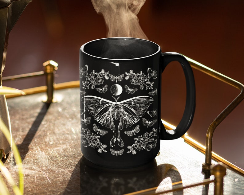 May include: Black ceramic mug featuring a white illustration of a moth, butterflies, flowers, and a crescent moon. The mug has a large handle and is filled with steaming liquid, suggesting a hot beverage.