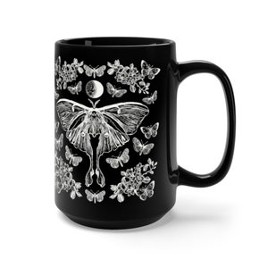May include: Black ceramic mug featuring a white illustration of a moth, butterflies, flowers, and a crescent moon. The mug has a large handle and a glossy finish. The design is symmetrical and detailed, creating a striking contrast against the dark background.