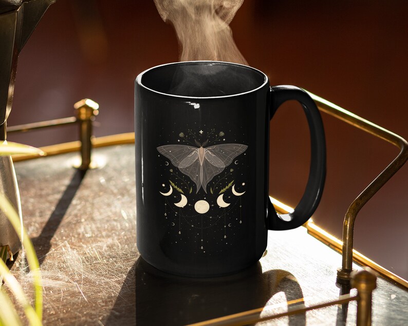 Pode incluir: Uma caneca de cer&acirc;mica preta com um design de mariposa e lua brancos. A caneca est&aacute; cheia de uma bebida fumegante e est&aacute; sobre uma bandeja dourada.