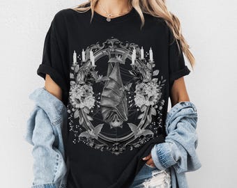 May include: Black t-shirt with a graphic of a bat hanging upside down in a decorative frame with candles and flowers. The bat is white and the frame is black.