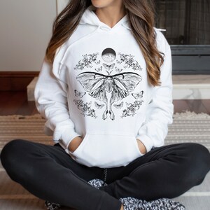 May include: White hoodie featuring a black and white graphic print of a moth with its wings outstretched. The moth is surrounded by smaller moths and floral designs. The graphic also includes a crescent moon.