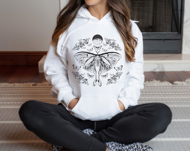 May include: White hoodie featuring a black and white graphic print of a moth with its wings outstretched. The moth is surrounded by smaller moths and floral designs. The graphic also includes a crescent moon.