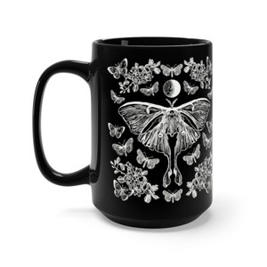 May include: Black ceramic mug featuring a white illustration of a moth, butterflies, flowers, and a moon phase design. The mug has a large handle and a glossy finish. The design is detailed and intricate, perfect for a nature lover.