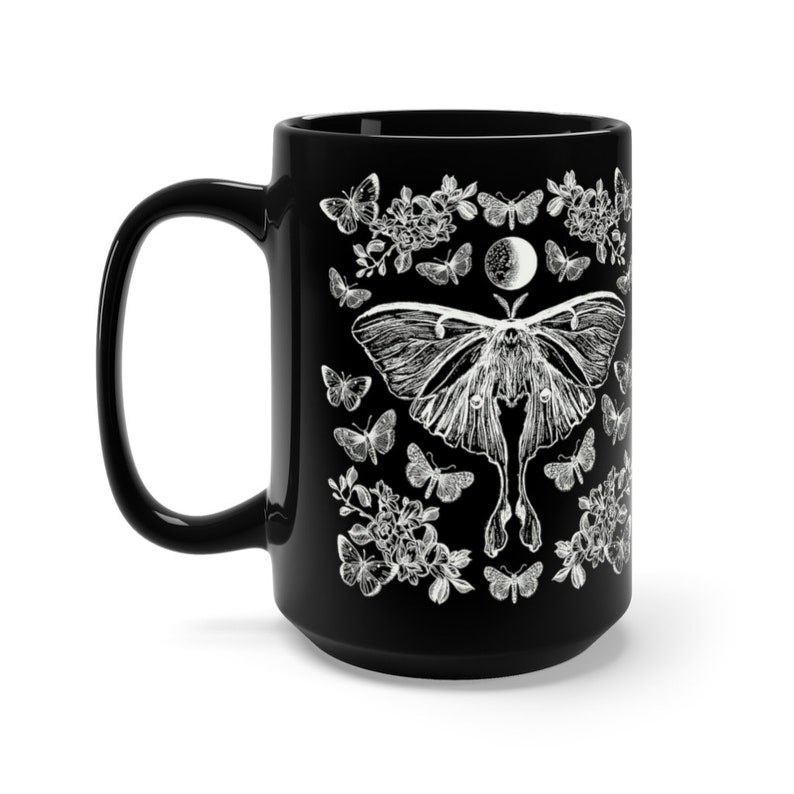 May include: Black ceramic mug featuring a white illustration of a moth, butterflies, flowers, and a moon phase design. The mug has a large handle and a glossy finish. The design is detailed and intricate, perfect for a nature lover.