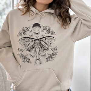 May include: A beige hoodie featuring a black and white graphic print of a moth, flowers, and a crescent moon.