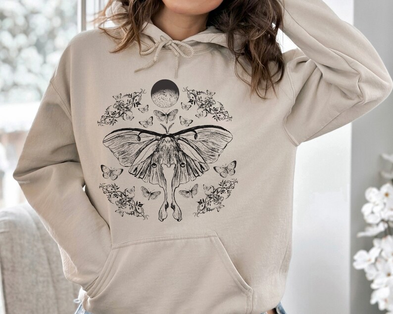 May include: A beige hoodie featuring a black and white graphic print of a moth, flowers, and a crescent moon.