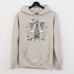 May include: A beige hoodie with a front pocket and a drawstring hood. The front of the hoodie features a black and white illustration of a bat hanging upside down from a branch, surrounded by flowers and candles. Two smaller bat silhouettes are also visible.