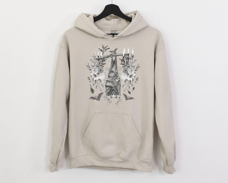 May include: A beige hoodie with a front pocket and a drawstring hood. The front of the hoodie features a black and white illustration of a bat hanging upside down from a branch, surrounded by flowers and candles. Two smaller bat silhouettes are also visible.