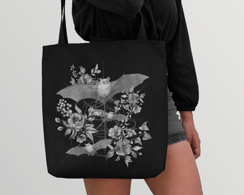 May include: Black tote bag with a white graphic design of bats and flowers. The bats are flying in a web-like pattern with flowers and leaves surrounding them.