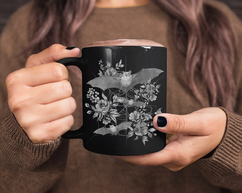 May include: Black ceramic mug with a white design of bats and flowers. The bats are flying in a circle around a spiderweb.