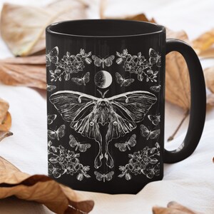 May include: Black ceramic mug featuring a detailed white illustration of a moth, butterflies, flowers, and a crescent moon. The design is set against a black background. The mug has a curved handle.