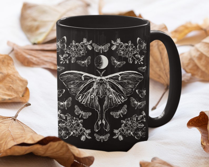 May include: Black ceramic mug featuring a detailed white illustration of a moth, butterflies, flowers, and a crescent moon. The design is set against a black background. The mug has a curved handle.