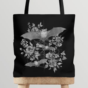 May include: Black tote bag with a white design of bats and flowers. The bats are flying in a web with flowers and leaves around them.